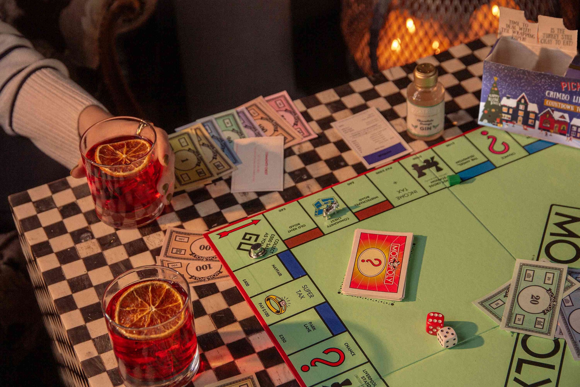 Person holding a drink next to a Monopoly game board with cards and dice on a checkered tablecloth.