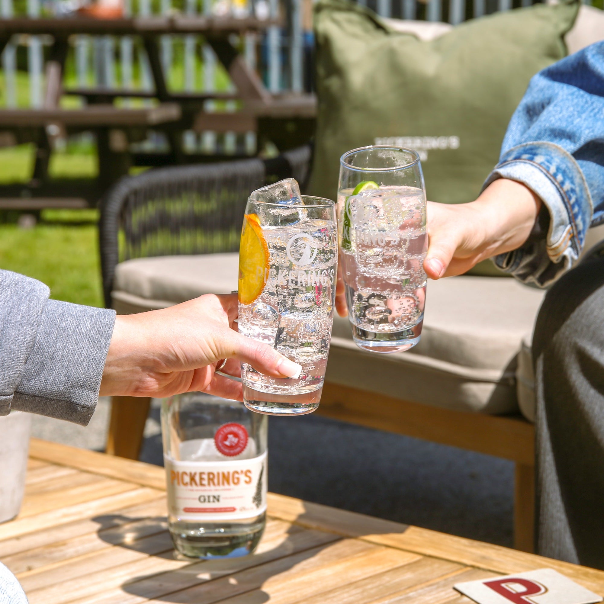 Two people sitting at a table outdoors, clinking glasses with Pickering's gin bottles.